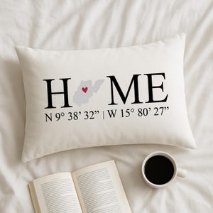 以下が含まれることがあります： クリーム色の長方形のクッションで、黒い大文字で「HOME」という文字が書かれています。「O」は赤いハートが付いたウェストバージニア州の地図に置き換えられています。その下に座標があります。コーヒーカップと開いた本も画像にあります。