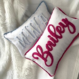 May include: Two white rectangular throw pillows with embroidered names. One pillow has the name "MINNA" in light blue, the other has "Bailey" in hot pink. Both pillows have a contrasting border.