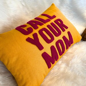 May include: A yellow throw pillow with the phrase "CALL YOUR MOM" embroidered in pink.