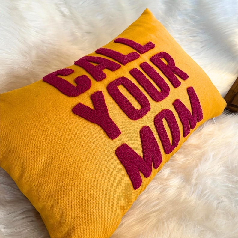 May include: A yellow throw pillow with the phrase "CALL YOUR MOM" embroidered in pink.