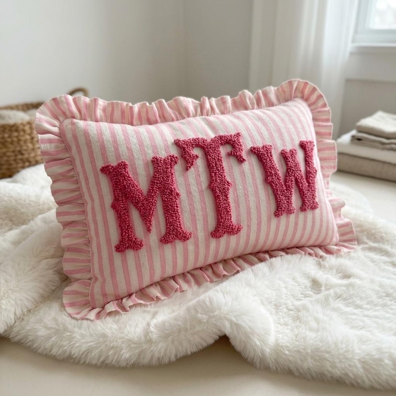 May include: A rectangular throw pillow featuring a pink and white striped pattern with a ruffled edge. The center of the pillow displays the letters MTW in large, textured, dark pink punch needle embroidery across the front surface.