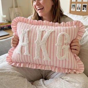 May include: A rectangular throw pillow featuring a light pink and white vertical striped pattern with a ruffled edge. The center of the pillow displays the letters AKG in large, white, textured, tufted fabric applique lettering.