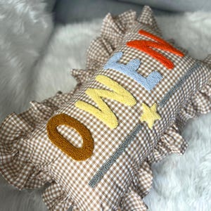 May include: A brown and white gingham pillow with the word "ONE" embroidered in orange, yellow, and blue. The pillow has a star and a decorative border. The pillow has a ruffled edge.