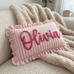 Pode incluir: Uma almofada retangular com padrão de riscas verticais em rosa claro e branco e uma borda com folhos. O nome Olivia está bordado numa fonte cursiva rosa-escura texturizada e em relevo no centro da superfície da almofada.