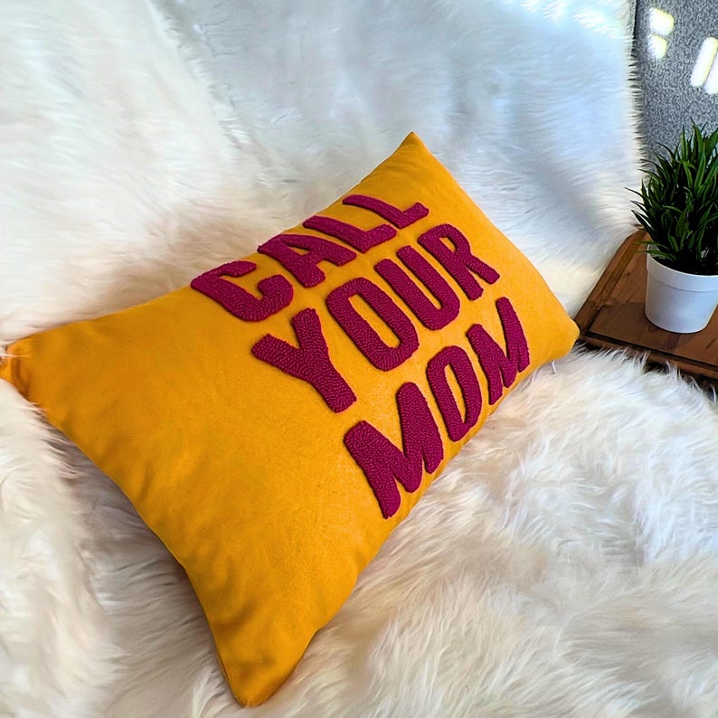 May include: A yellow throw pillow with the phrase "CALL YOUR MOM" embroidered in pink.