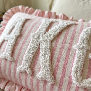 May include: A rectangular throw pillow featuring a light pink and white vertical striped fabric base with a ruffled edge. The surface displays large, white, textured, punch needle embroidered letters that spell out the name AKYLA.