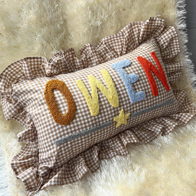 May include: A rectangular pillow with a brown and white gingham pattern and a ruffled edge. The word "OWEN" is embroidered in orange, yellow, blue, and red. A yellow star is embroidered below the "W".