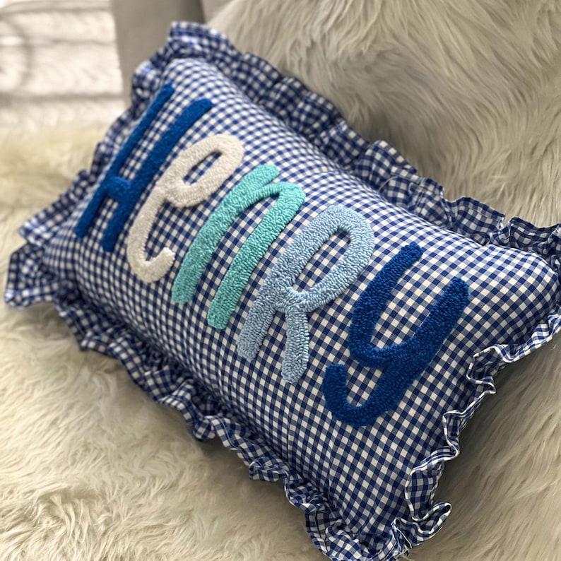 May include: A rectangular pillow with a blue and white gingham pattern. The word "HENRY" is embroidered across the pillow in various shades of blue and white. The pillow has a ruffled edge.