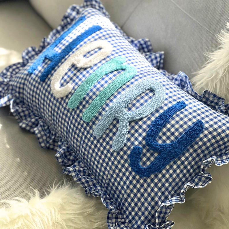 May include: A rectangular pillow with a blue and white gingham pattern. The word "HAPPY" is embroidered in varying shades of blue and white. The pillow has a ruffled edge.