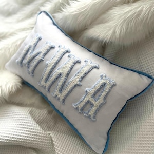May include: A rectangular white pillow with the embroidered word "MAMA" in light blue and white. The pillow has a light blue border and is resting on a white, textured surface.