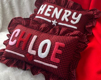 May include: Two red and black gingham pillows with white embroidered names "HENRY" and "CHLOE". The pillows have ruffled edges and are on a white fluffy surface with a red blanket.