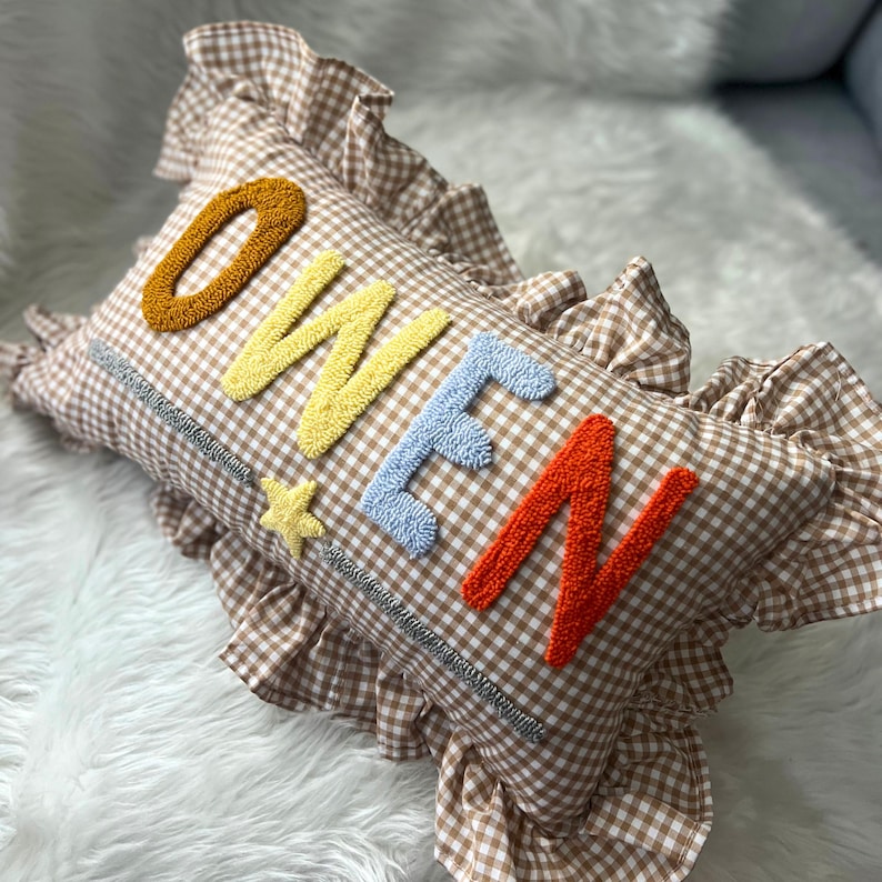May include: A rectangular pillow with a brown and white gingham pattern. The word "OWEN" is embroidered in colorful, textured letters: gold, yellow, blue, and orange. A yellow star is embroidered next to the "O". The pillow has a ruffled edge.