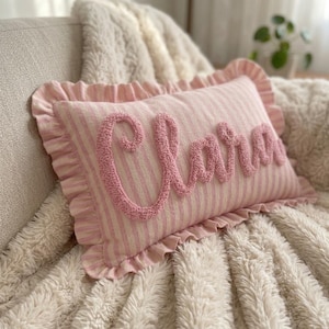 May include: A rectangular throw pillow featuring a light pink and white vertical striped pattern. The pillow has a ruffled border and the name Clara embroidered in a raised, textured pink script font across the center of the front surface.