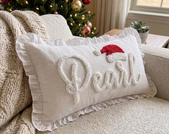 May include: A rectangular off-white throw pillow with a ruffled edge, featuring the name Pearl embroidered in white script. A small red and white Santa hat is positioned above the letter P. The pillow has a textured, fabric-like appearance.