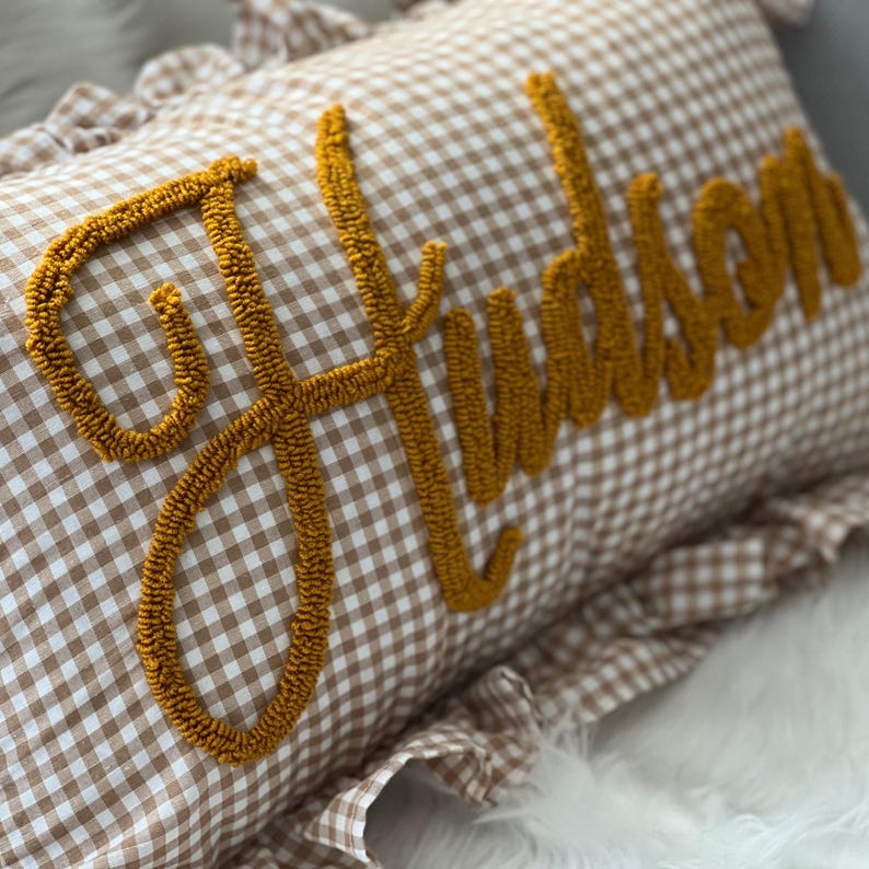 K&ouml;nnte beinhalten: Ein dekoratives Kissen mit braun-wei&szlig;em Vichy-Muster. Der Name "Hudson" ist in senfgelber, strukturierter Schrift auf die Vorderseite gestickt. Das Kissen hat einen ger&uuml;schten Rand.