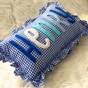 May include: A blue and white gingham pillow with the word "HOME" embroidered in varying shades of blue and white. The pillow has a ruffled edge, adding a decorative touch to the home decor item.