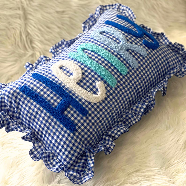 May include: A blue and white gingham pillow with the word "HOME" embroidered in varying shades of blue and white. The pillow has a ruffled edge, adding a decorative touch to the home decor item.