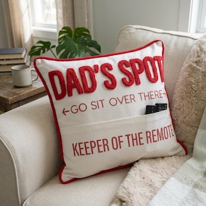 May include: Cream-colored decorative pillow with red trim and text. The pillow reads "DAD'S SPOT" in raised red letters, with "GO SIT OVER THERE" and "KEEPER OF THE REMOTE" printed below. A pocket holds two remote controls.