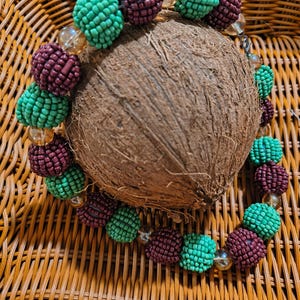 May include: A beaded bracelet with alternating maroon and teal beads, interspersed with clear beads. The bracelet is resting on a brown coconut, set against a woven wicker background. The beads have a textured, clustered appearance.