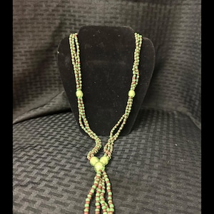 Puede incluir: Un collar de múltiples hilos con pequeñas cuentas alternas de color verde lima y marrón rojizo. El diseño incluye tres cuentas esféricas más grandes y texturizadas de color verde lima, colocadas simétricamente con un nudo central.