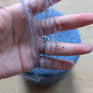May include: A hand displaying strands of light blue yarn, with a ball of yarn behind. The yarn looks soft and fluffy, suitable for knitting or other crafts. The colour is a muted, dusty blue.