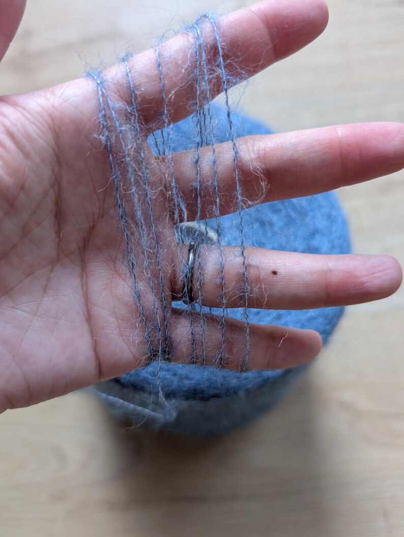 May include: A hand displaying strands of light blue yarn, with a ball of yarn behind. The yarn looks soft and fluffy, suitable for knitting or other crafts. The colour is a muted, dusty blue.