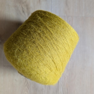 May include: A large spool of yellow-green yarn. The yarn is tightly wound and has a slightly fuzzy texture. The spool is angled, showing the layers of yarn. The background is a light wood surface.