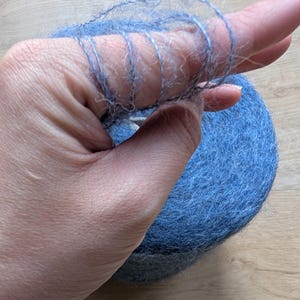 May include: A ball of blue yarn with a hand holding a strand of the yarn. The yarn is a soft, fuzzy material. The hand is holding a small section of the yarn, showing the texture and colour.