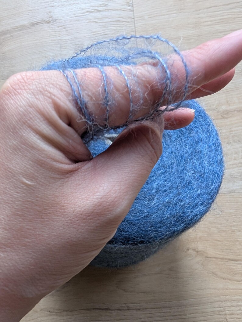 May include: A ball of blue yarn with a hand holding a strand of the yarn. The yarn is a soft, fuzzy material. The hand is holding a small section of the yarn, showing the texture and colour.