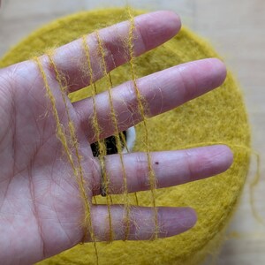 May include: Close-up of a skein of yellow yarn with strands held between fingers. The yarn is a vibrant yellow color, with a soft, fuzzy texture. The skein is round and appears to be made of a natural fiber, perfect for knitting or crocheting projects.