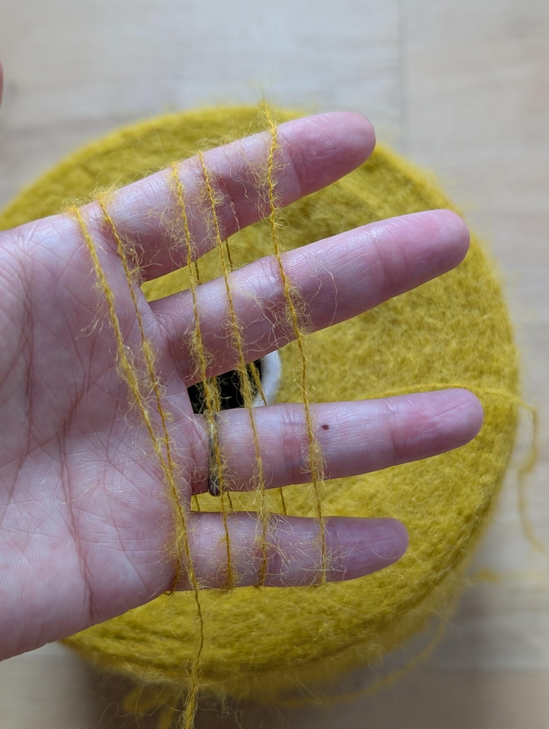 May include: Close-up of a skein of yellow yarn with strands held between fingers. The yarn is a vibrant yellow color, with a soft, fuzzy texture. The skein is round and appears to be made of a natural fiber, perfect for knitting or crocheting projects.