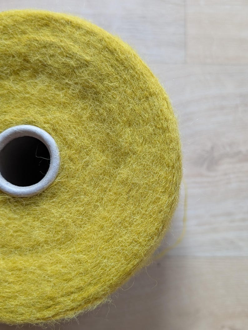 May include: Close-up of a large spool of yellow yarn. The yarn is a vibrant, solid color and appears soft and textured. The spool has a white cardboard core. The yarn is likely used for knitting or crocheting projects.