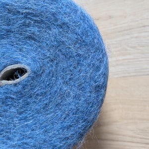 May include: A tightly wound roll of blue yarn, showing a cardboard core. The yarn has a soft, textured appearance, possibly wool or a similar fibre. The colour is a medium shade of blue.