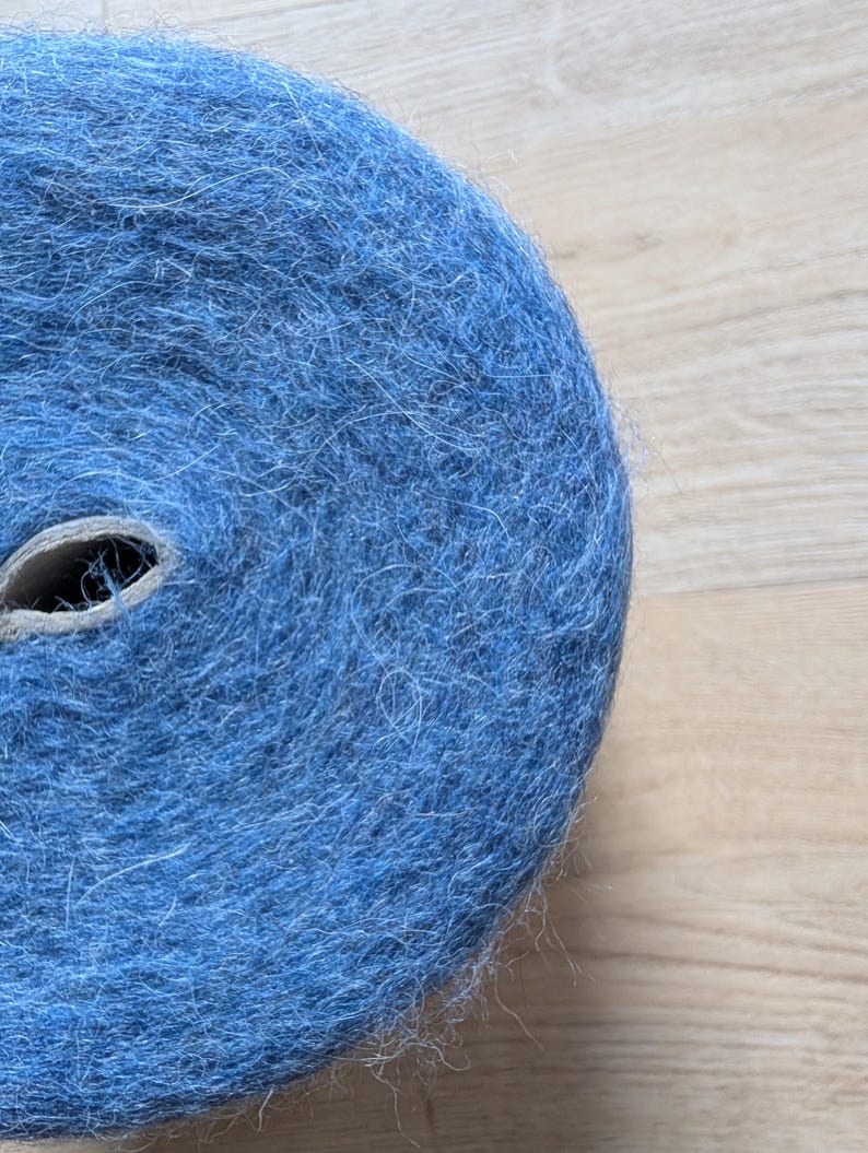 May include: A tightly wound roll of blue yarn, showing a cardboard core. The yarn has a soft, textured appearance, possibly wool or a similar fibre. The colour is a medium shade of blue.