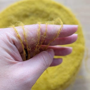 May include: Close-up of a hand holding strands of yellow yarn. The yarn is a vibrant mustard color and appears soft and fluffy. A large, circular bundle of the same yarn is in the background, suggesting a craft or textile project.