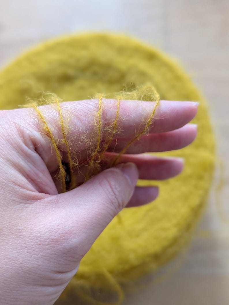 May include: Close-up of a hand holding strands of yellow yarn. The yarn is a vibrant mustard color and appears soft and fluffy. A large, circular bundle of the same yarn is in the background, suggesting a craft or textile project.