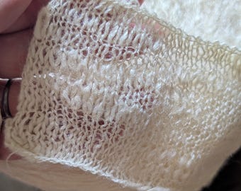 May include: Close-up of a cream-colored, open-weave knit fabric. The fabric has a delicate, airy texture with visible stitches and small holes. The edge of the fabric is visible, showing a finished, slightly ruffled border. The overall appearance is soft and lightweight.