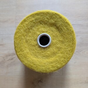 May include: Overhead view of a large spool of yellow yarn. The yarn is a vibrant, mustard yellow color and appears soft and fluffy. The spool has a white cardboard core with a black center hole. The yarn is likely for knitting or crocheting projects.