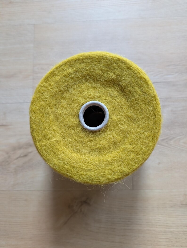 May include: Overhead view of a large spool of yellow yarn. The yarn is a vibrant, mustard yellow color and appears soft and fluffy. The spool has a white cardboard core with a black center hole. The yarn is likely for knitting or crocheting projects.