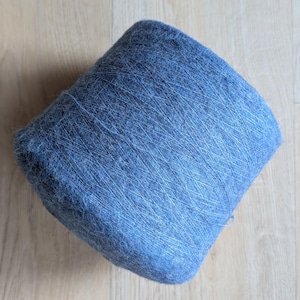 May include: A large, tightly wound skein of blue yarn. The yarn appears soft and fluffy, with a slightly textured surface. The colour is a muted, dusty blue, and the yarn is neatly coiled. The background is a light-coloured wooden surface.