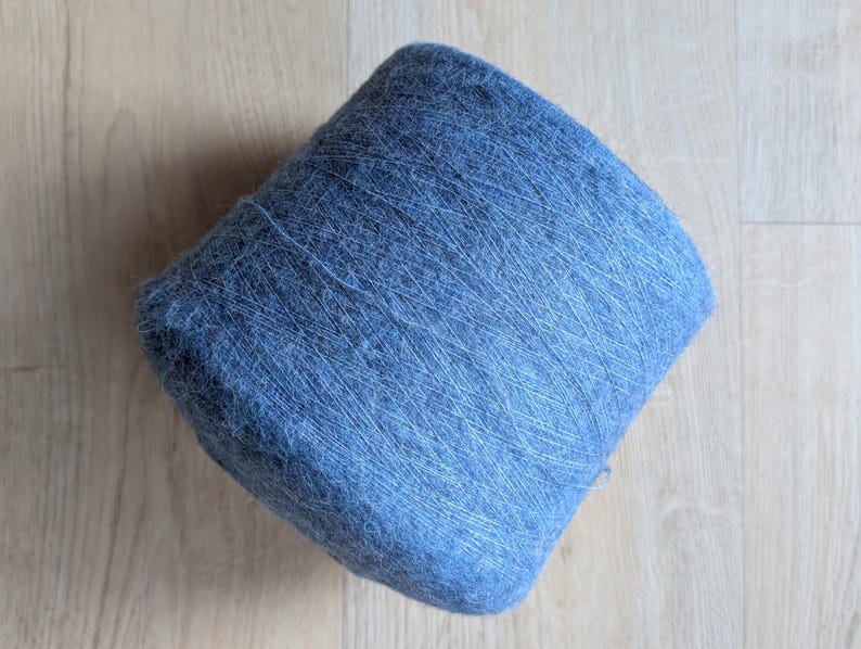 May include: A large, tightly wound skein of blue yarn. The yarn appears soft and fluffy, with a slightly textured surface. The colour is a muted, dusty blue, and the yarn is neatly coiled. The background is a light-coloured wooden surface.