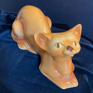 May include: A glossy, orange ceramic cat figurine in a seated, resting position. The cat features large, pointed ears, almond-shaped yellow eyes, a small brown nose, and subtle whiskers painted on its face. The smooth, glazed surface reflects light.