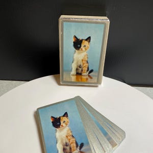 May include: A deck of playing cards featuring a calico kitten on the back of each card. The cards are displayed in a fanned arrangement alongside the matching rectangular card box, both showcasing the same kitten image against a blue background.