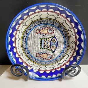 May include: A round ceramic bowl featuring a central mosaic-style design of three fish in yellow, orange, and blue. The rim is decorated with a bold blue wavy pattern, while the inner border displays intricate black, white, and blue geometric motifs.