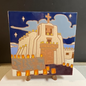 May include: A square ceramic tile featuring a colorful illustration of an adobe-style church at night. The scene depicts a cream-colored building with a cross, illuminated by numerous small orange lights against a dark blue sky with stars.