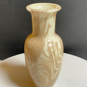 May include: A glass vase featuring a swirling marbled pattern of cream and light brown colors. The vase has a rounded body, a narrow neck, and a flared rim. The surface has a smooth, glossy finish that reflects light, creating a polished appearance.
