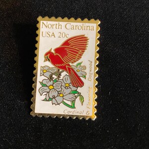 Pu&ograve; includere: Spilla con bordo dorato a forma di francobollo. Il francobollo raffigura un uccello cardinale rosso posato su fiori di corniolo bianchi e grigi. Il testo sul francobollo recita "North Carolina USA 20c" e "Cardinal & Flowering Dogwood."