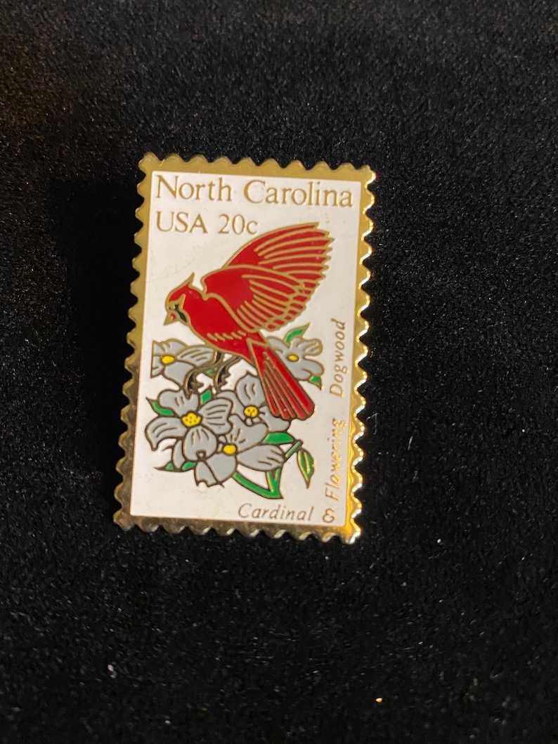 Pu&ograve; includere: Spilla con bordo dorato a forma di francobollo. Il francobollo raffigura un uccello cardinale rosso posato su fiori di corniolo bianchi e grigi. Il testo sul francobollo recita "North Carolina USA 20c" e "Cardinal & Flowering Dogwood."