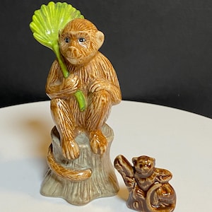 May include: A ceramic monkey figurine sitting on a tree stump and holding a bright green leaf, accompanied by a smaller, separate brown monkey figurine. Both figures feature glossy brown finishes with detailed textures and painted facial features.