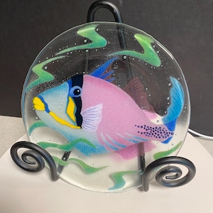 May include: A circular fused glass plate featuring a vibrant triggerfish with a pink body, blue fins, and yellow accents. The fish is surrounded by abstract green wavy lines on a clear glass background, displayed on a black metal stand.